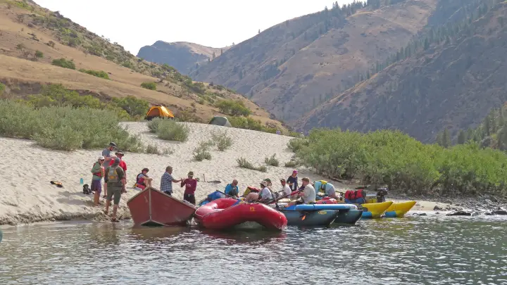 A large group unloading rafts onto a wide sand beach beneath grassy hills